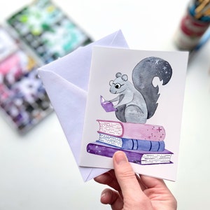 May include: A watercolor illustration of a gray squirrel wearing glasses and reading a book while sitting on a stack of three books. The books are purple, blue, and pink with a white background.