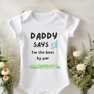 May include: White baby bodysuit with the text "Daddy Says I'm the best by par" in black. The design features a golf flag and green grass. The bodysuit is suspended from a wooden clip.