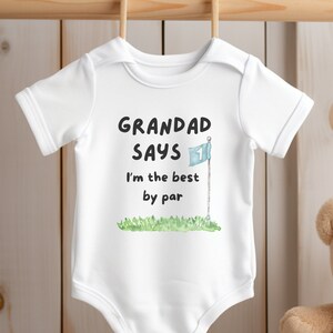 May include: White baby onesie with the text "GRANDAD SAYS I'm the best by par" in black. A golf flag graphic is included. The onesie is hanging on a wooden rod. A teddy bear is in the background.