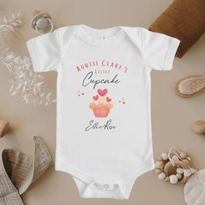 May include: White baby onesie with the text "Auntie Clare's Little Cupcake" and a pink cupcake with red hearts on top. The name "Ellie-Rose" is written below the cupcake.