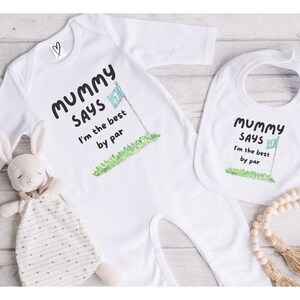 May include: White baby onesie and bib set with the text "Mummy Says I'm the best by par" in black. The design includes a golf flag illustration. A stuffed bunny toy is also in the image.