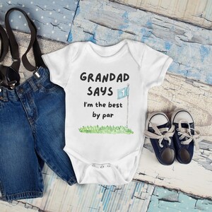 May include: White baby onesie with the text "GRANDAD SAYS I'm the best by par" and a golf flag graphic. The onesie is next to a pair of blue denim shorts with suspenders and navy blue baby shoes.