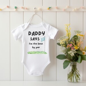 May include: White baby onesie with the text "DADDY SAYS I'm the best by par" in black font. A watercolor illustration of a golf flag and green grass is below the text. The onesie is hanging on a white wall.