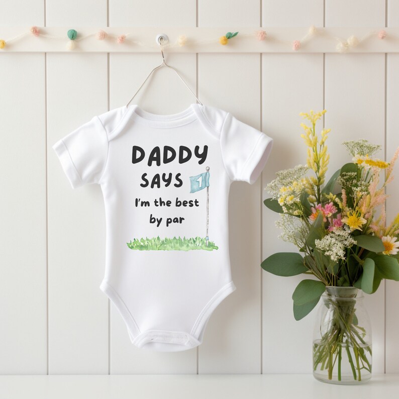 May include: White baby onesie with the text "DADDY SAYS I'm the best by par" in black font. A watercolor illustration of a golf flag and green grass is below the text. The onesie is hanging on a white wall.