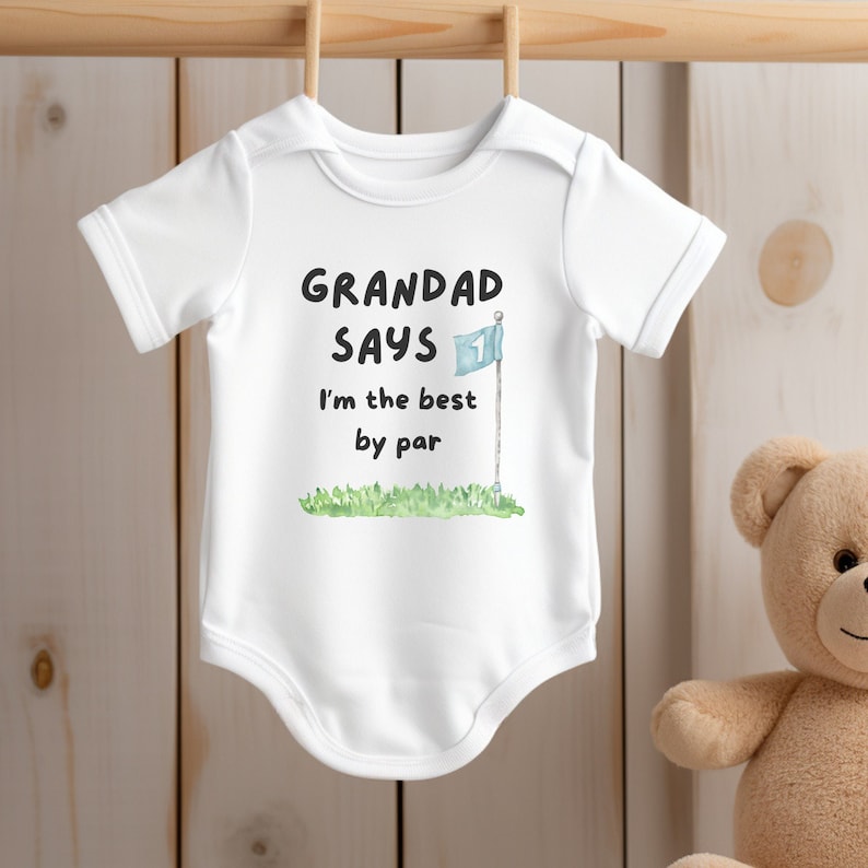 May include: White baby onesie with the text "GRANDAD SAYS I'm the best by par" in black. A golf flag graphic is included. The onesie is hanging on a wooden rod. A teddy bear is in the background.
