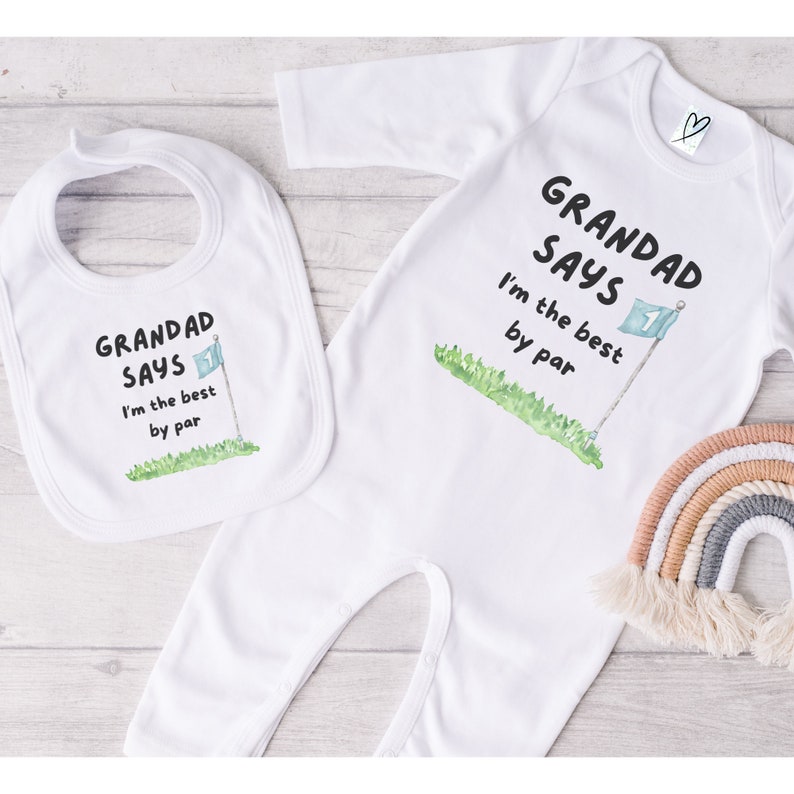 May include: White baby bib and onesie set. The bib and onesie both feature the text "GRANDAD SAYS I'm the best by par" with a golf flag graphic. A small rainbow decorates the background.