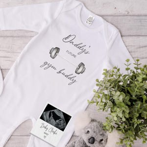 Pode incluir: Um macacão branco para bebê com o texto "Daddy's Future Gym Buddy" e uma ilustração em preto e branco de uma barra de halteres. Uma imagem de ultrassom em preto e branco está presa ao macacão com o texto "Baby Clarke 2027".