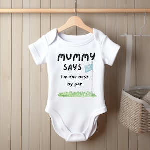 May include: White baby onesie with the text "Mummy Says I'm the best by par" in black. A watercolor illustration of a golf flag and green grass is below the text. The onesie is hanging on a wooden hanger.