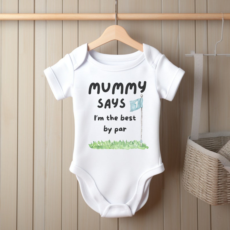 May include: White baby onesie with the text "Mummy Says I'm the best by par" in black. A watercolor illustration of a golf flag and green grass is below the text. The onesie is hanging on a wooden hanger.