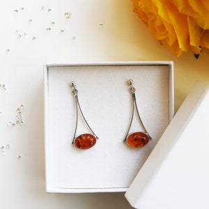 Könnte beinhalten: Ein Paar silberne Ohrhänger mit ovalen Bernstein-Steinen, präsentiert in einer weißen Box mit dem Text "FILIGREE STREET".