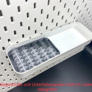 May include: White plastic storage tray with a gray compartment for holding bobbins. The tray is mounted on a white pegboard. The text "Ikea Ablageschale und Unterfadenspulen nicht im Lieferumfang inbegriffen" is visible at the bottom of the image.