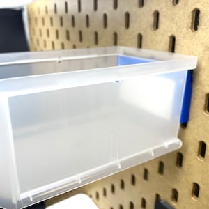 May include: A clear plastic storage bin with a blue accent, mounted on a pegboard. The bin has a rectangular shape with curved sides and is designed for organizing small items. The pegboard is a light brown colour.