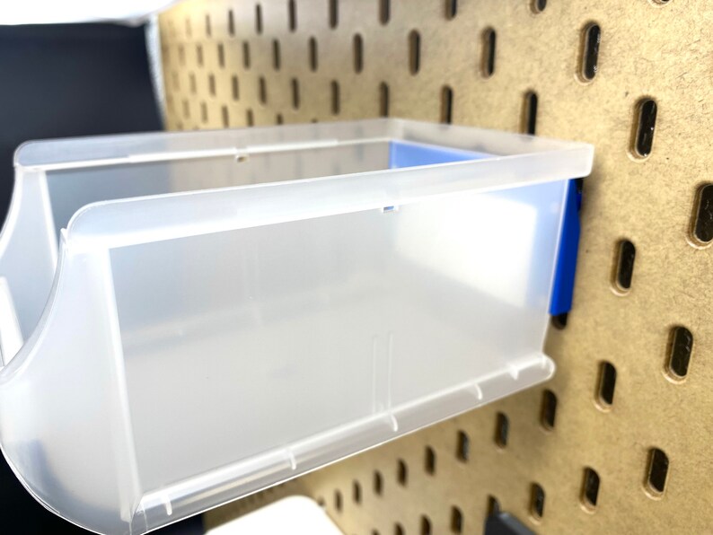 May include: A clear plastic storage bin with a blue accent, mounted on a pegboard. The bin has a rectangular shape with curved sides and is designed for organizing small items. The pegboard is a light brown colour.