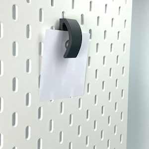 Könnte beinhalten: Ein schwarzer Plastikhaken hängt an einer weißen Lochwand, an dem ein weißes Blatt Papier hängt.