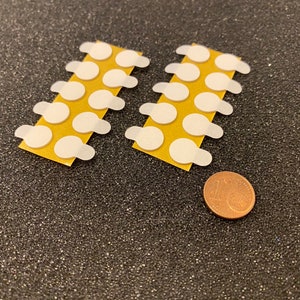 May include: Two strips of adhesive dots, each with 10 white, round adhesive dots on a brown backing. A copper-colored coin is below the strips for size reference.