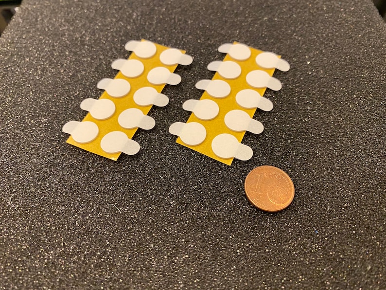 May include: Two strips of adhesive dots, each with 10 white, round adhesive dots on a brown backing. A copper-colored coin is below the strips for size reference.