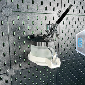 Könnte beinhalten: Eine Airbrush mit schwarzem Griff und silberner Düse ist an einer dunkelgrauen Lochwand befestigt. Die Airbrush ist an einem transparenten, zylindrischen Farbbehälter befestigt, der von einer weißen Halterung gehalten wird. Die Lochwand hat zahlreiche Löcher.