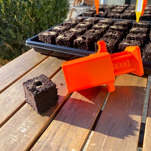 Könnte beinhalten: Ein orangefarbenes Plastikwerkzeug zum Pflanzen von Saatgut-Startwürfeln. Das Werkzeug hat einen Griff und einen Schlitz, um den Würfel zu halten. Ein brauner Saatgut-Startwürfel liegt auf einer Holzoberfläche.