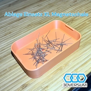 May include: An orange rectangular tray with a collection of silver pins and a single red pin. The tray has the text "Ablage Einsatz XL Magnetschale" and the logo "3DVERSUM". The tray is on a wooden surface.