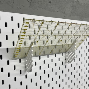 Può includere: Un pannello forato bianco con un righello di plastica trasparente appeso a due ganci bianchi. Il righello è graduato in pollici e centimetri.