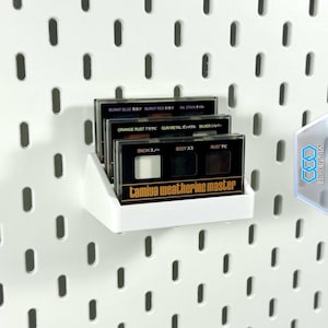 Könnte beinhalten: Ein weißes Regal hält mehrere Tamiya Weathering Master-Kits vor einer weißen Lochwand. Die Kits sind schwarz mit Farbbezeichnungen auf Japanisch und Englisch, darunter "Burnt Blue", "Burnt Red" und "Oil Stain". Das Regal ist an einer weißen Lochwand befestigt.