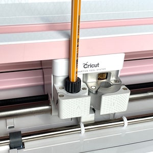 Könnte beinhalten: Nahaufnahme einer Cricut Explore Air 2 Schneidemaschine mit einem gelben Bleistift, der in den Werkzeughalter eingesetzt ist. Die Maschine hat ein rosa-weißes Design und der Text "Cricut Adaptive Tool System" ist sichtbar.