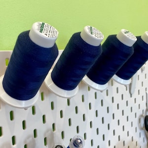 Può includere: Quattro bobine di filo blu scuro sono appese a una bacheca bianca. Le bobine sono etichettate "Wonderfil".