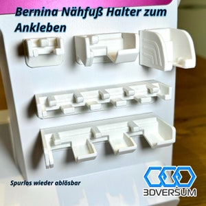 Könnte beinhalten: Weißer Kunststoff-Nähfußhalter mit mehreren Schlitzen zum Verstauen von Nähfüßen. Der Halter ist so konzipiert, dass er mit Klebstoff an einer Nähmaschine befestigt werden kann. Der Text "Bernina Nähfuß Halter zum Ankleben" und "Spurlos wieder ablösbar" ist auf dem Halter aufgedruckt. Der Text "3DVERSUM" ist in der unteren rechten Ecke des Halters aufgedruckt.