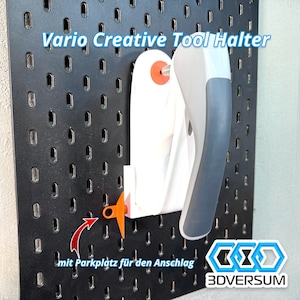 Può includere: Portautensili in plastica bianca con manico grigio, montato su una bacheca nera. Il portautensili ha una piccola linguetta arancione che può essere utilizzata per regolare la posizione dell'utensile. Il testo "Vario Creative Tool Halter" è stampato sulla bacheca. Il testo "mit Parkplatz für den Anschluss" è stampato sotto il portautensili. Il logo "3DVERSUM" è stampato nell'angolo in basso a destra.