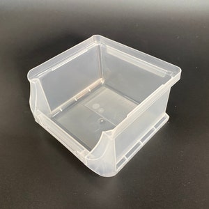 Può includere: Un contenitore di stoccaggio in plastica trasparente con forma quadrata e un bordo attorno al bordo superiore. Il contenitore è vuoto e si trova su una superficie nera.