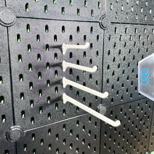 Op de afbeelding: Een zwart pegboard met meerdere rijen gelijkmatig verdeelde gaten. Vier gebroken witte, gebogen haken zijn in het bord geplaatst. Het pegboard is gemaakt van een mat materiaal. De achtergrond heeft een groene tint.