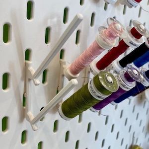 Könnte beinhalten: Eine weiße Lochwand mit mehreren Garnrollen in verschiedenen Farben, darunter rosa, rot, blau, grün und lila. Die Rollen werden von weißen Kunststoffhaltern gehalten. Die Lochwand hat ein Raster aus Löchern zum Aufhängen von Gegenständen.