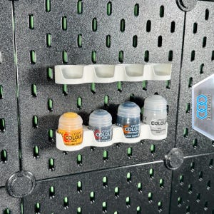 Könnte beinhalten: Weiße Kunststoff-Farbtopfhalterungen, montiert auf einer schwarzen Lochplatte. Die Halterungen sind für Citadel Colour Farbtöpfe konzipiert. Das Bild zeigt mehrere Farbtöpfe in verschiedenen Farben, darunter Gelb, Blau und Grau.