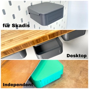 May include: Three gray plastic storage containers, two with lids, one without. The container without a lid is labeled "Desktop". The container with a lid is labeled "für Skadis". The third container is on a brown surface and is labeled "Independent".