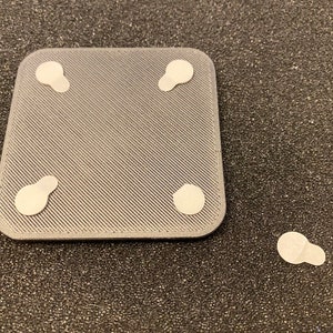 May include: A gray square with four white adhesive pads on the bottom. The pads are slightly oval shaped.