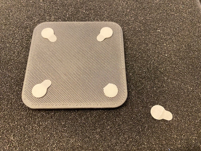 May include: A gray square with four white adhesive pads on the bottom. The pads are slightly oval shaped.