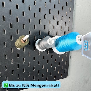 Op de afbeelding: Een zwart pegboard met verticale sleuven met drie klossen garen in olijfgroen, grijs en felblauw. Een wit plastic label met de blauwe tekst 3DVERSUM is bevestigd aan de blauwe klos. Een lichtblauwe banner toont de tekst Bis zu 15% Mengenrabatt.