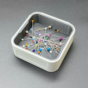 Può includere: Un cuscino quadrato bianco pieno di una varietà di spilli colorati. Gli spilli hanno teste di diversi colori, tra cui bianco, blu, arancione e rosa. Il cuscino ha un interno in rete grigia.