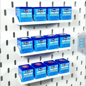 Könnte beinhalten: Eine Präsentation von Revell Aqua Color Acrylfarben in kleinen blauen Boxen, die jeweils 18 ml Farbe enthalten. Die Boxen sind auf weißen Regalen vor einer weißen Lochwand angeordnet und mit dem Markennamen versehen.