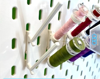 Può includere: Un gancio in plastica bianca per pannelli forati con due punte angolate che sostengono diverse bobine di filo da cucito in vari colori, tra cui rosa chiaro, rosso scuro, verde oliva e viola. Il pannello presenta una griglia di fessure.