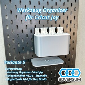 May include: A gray and white tool organizer for Cricut Joy, mounted on a black pegboard. The organizer has multiple compartments and includes a magnetic storage tray. Text on the image reads "Werkzeug Organizer f&uuml;r Cricut Joy" and "Variante 5".