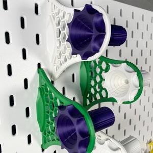 K&ouml;nnte beinhalten: Drei 3D-gedruckte Lochwand-Organizer in Wei&szlig;, Gr&uuml;n und Lila. Die Organizer haben ein Wabenmuster und sind an einer wei&szlig;en Lochwand befestigt. Die Organizer sind f&uuml;r die Aufnahme verschiedener Gegenst&auml;nde konzipiert.