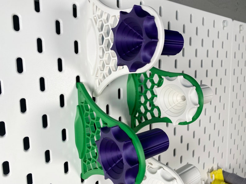 K&ouml;nnte beinhalten: Drei 3D-gedruckte Lochwand-Organizer in Wei&szlig;, Gr&uuml;n und Lila. Die Organizer haben ein Wabenmuster und sind an einer wei&szlig;en Lochwand befestigt. Die Organizer sind f&uuml;r die Aufnahme verschiedener Gegenst&auml;nde konzipiert.