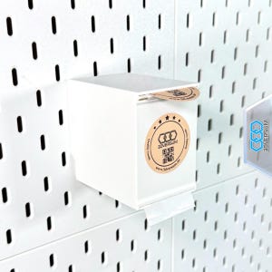 Op de afbeelding: Een witte, rechthoekige dispenserbox bevestigd aan een wit pegboard. De doos heeft een ronde sticker met de tekst "3DVERSUM" en een QR-code. Een klein stukje papier wordt afgegeven. Het pegboard heeft gelijkmatig verdeelde sleuven.