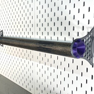 K&ouml;nnte beinhalten: Ein zylindrisches Objekt mit schwarzem und goldenem Muster, m&ouml;glicherweise ein Werkzeug oder Zubeh&ouml;r, ist mit grauen und violetten Haltern an einer wei&szlig;en Lochwand befestigt. Die Halter haben ein geometrisches Design. Das Objekt ist zylindrisch und hat eine strukturierte Oberfl&auml;che.