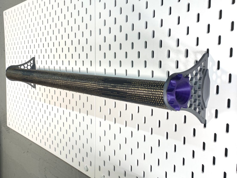 K&ouml;nnte beinhalten: Ein zylindrisches Objekt mit schwarzem und goldenem Muster, m&ouml;glicherweise ein Werkzeug oder Zubeh&ouml;r, ist mit grauen und violetten Haltern an einer wei&szlig;en Lochwand befestigt. Die Halter haben ein geometrisches Design. Das Objekt ist zylindrisch und hat eine strukturierte Oberfl&auml;che.