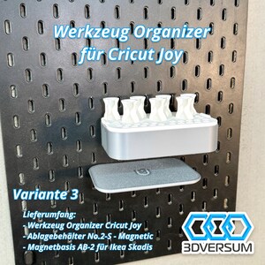 May include: A gray and white organizer for Cricut Joy tools, mounted on a black pegboard. The organizer has multiple slots for tools and a lower shelf. Text on the image reads "Werkzeug Organizer f&uuml;r Cricut Joy" and "Variante 3".