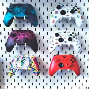 Può includere: Sei diversi controller per videogiochi in vari colori e design. I controller sono appesi a una parete bianca con pannelli forati. I controller sono tutti di marchi e modelli diversi.
