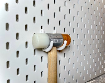 Könnte beinhalten: Eine weiße Lochwand mit einem weißen und gelben Gummihammer, der daran hängt. Der Hammer hat einen weißen Kopf und einen Holzstiel. Der Hammer hängt an zwei weißen Haken. Auf dem Hammer steht "GERMANY" gedruckt.