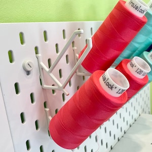 Può includere: Una lavagna forata bianca con diversi rocchetti di filo di vari colori, tra cui rosso, verde acqua e blu. I rocchetti di filo sono etichettati con il marchio "Trojalock" e il peso del filo "120". La lavagna forata ha un supporto in plastica bianca per i rocchetti.